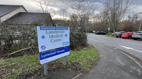A white and blue sign that reads 'Lanchester Medical Centre" in a grey car park area.