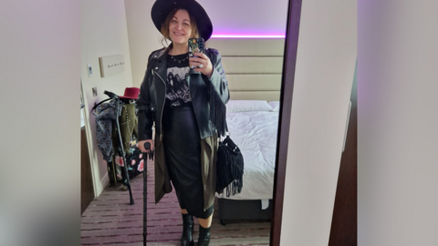 A woman in a black leather jacket, hat, dress and boots. She is standing with a walking stick. 