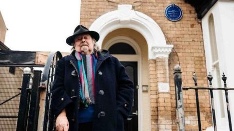 A man is stood outside an open black gate which leads to the front door of a property with an archway. He is wearing a thick black coat, black hat and colourful scarf.
