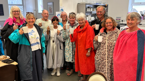 A group of 10 people wearing dryrobes or wraps which are used after swimming to keep warm, all holding white coffee mugs and smiling in an indoor cafe area