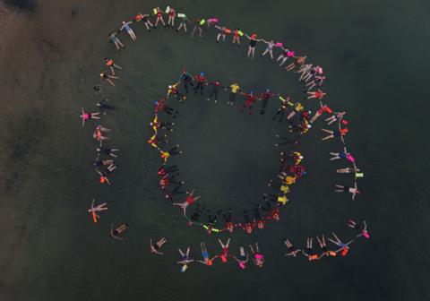 a group of swimmers floating together in a circle photographed from the air many are wearing brightly coloured clothes and holding hands
