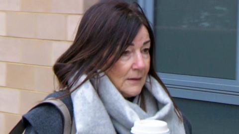 A woman with dark straight hair. She looks cold, and is wearing a grey designer scarf. She is carrying a costa cup. 