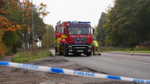 Images shows a red fire and rescue engine parked on a road. There are fire crew members at both sides of the truck wearing high-visibility uniforms, they are looking inside the fire engine. In the foreground, just in front of where the photo has been take, is police tape. 