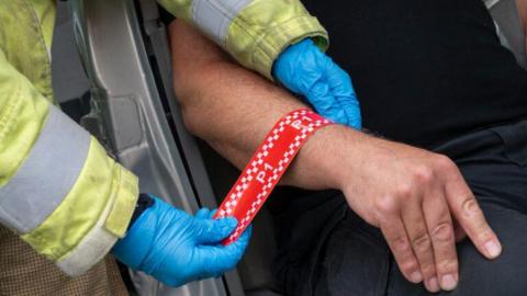 A triage band, in red, with P1, written on it, being put on a mans hand,  by a firefighter. The man wearing dark clothing is sat in a car.