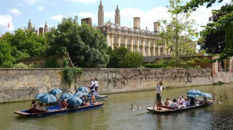 Punting in Cambridge. Three punts can be seen next to a walled section of the river. A university college building is in the background, and the spires or King's College Chapel.