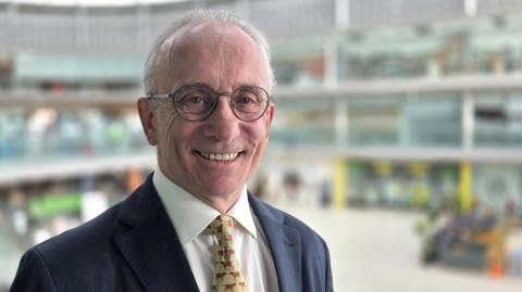 David Bick is smiling. He's wearing a navy suit jacket over a white shirt and patterned tie. The inside of the Norwich Forum is behind him but out of focus.