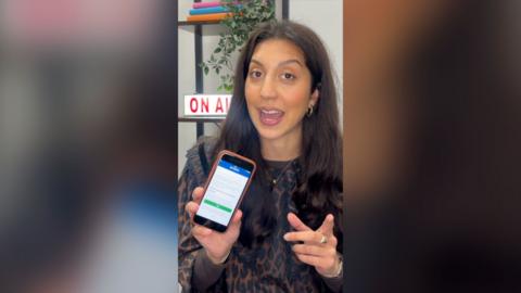 A woman with brown hair holds up an iPhone showing an app.