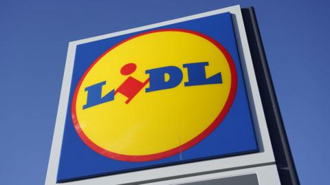 The image shows a Lidl logo on a tall sign. It is a blue square, with a yellow circle outlined in red. The word "Lidl" is written in blue font and the I is red. The sky is blue behind the sign.