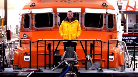 This is a photo of Richard Evans standing in front of a bright orange lifeboat. He is wearing a yellow waterproof jacket and black trousers.