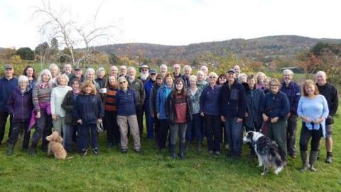 A large group of people in outdoor clothing stand in a field with two dogs and grassy hills behind them.