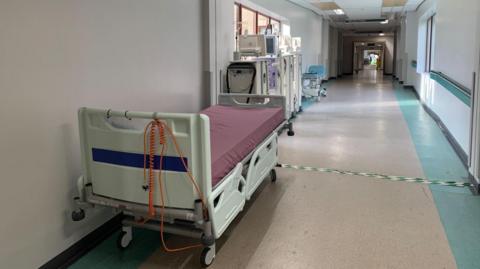 An empty portable hospital bed is at the side of a long empty hospital corridor. There are portable stands behind it with monitors and equipment.