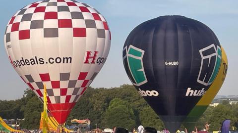 Generic shot of two tethered hot air balloons in a park.
One balloon is white with red and grey checks.  The other is navy blue with a corporate logo.