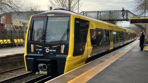 A Merseyrail black and yellow electric train on the track. A bridge over the platform can be seen above the train with a man walking across the other side. One man can be seen standing on the train platform at the bottom looking towards the train.