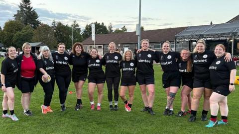 The Galts line up for a team photo. They are standing on a rugby pitch, all wearing a black kit, and they have their arms around each other. The clubhouse can be seen behind them.