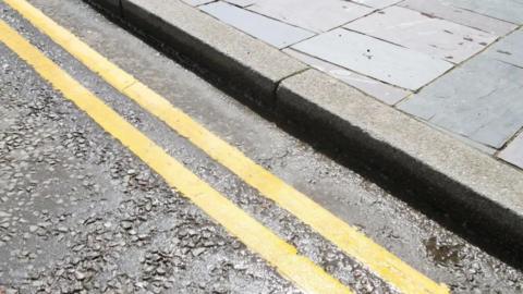 A close-up shot of double yellow lines on a road, next to a kerb and a pavement.