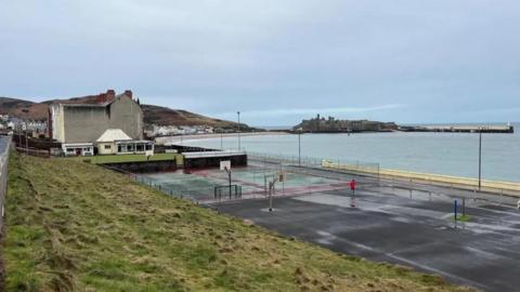 Unkept sports court opposite the sea with Peel Castle in the distance.