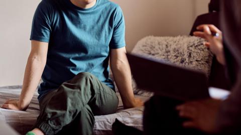 Down syndrome man sitting on sofa during conversation with a psychologist. He is wearing a blue top and green trousers, the person to the left, has an arm out, but is blurred. 