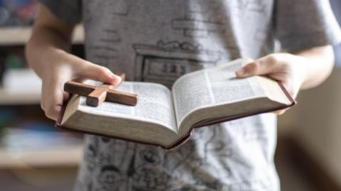 Someone holds a book in their hands with brown edging, while holding a brown cross in their right hand.