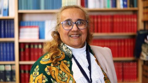 Gulshan Kayembe wearing a long Kashmiri embroidered jacket and the High Sheriff pendant. She standing in front of rows of book shelves.