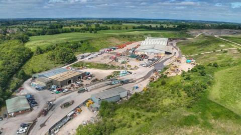 An aerial view of the quarry, surrounding by green fields. Numerous buildings separated by roads and car parks can be seen.