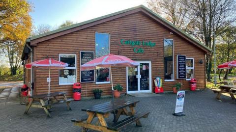 Cartgate Lodge Cafe. It is a large wooden clad shed with tall windows. There are three picnic benches and parasols outside on the paved area, and trees dotted around.