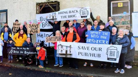 Members of the Brydekirk community gathered in front of the former pub holding an array of banners titled with slogans such as 'save our pub/hub'.
