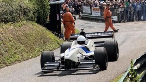 A white formula 1 style racing car on a tarmac track bordered by a grassy verge and embankment. Crowds stand behind a railing at the side of the track and lean over barriers at the side while crew in orange jumpsuits walk across it behind the vehicle.