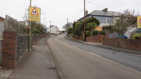 Marldon Road in Paignton. Yellow signs saying "CAUTION Pedestrians crossing" are on signposts. It is an overcast day.