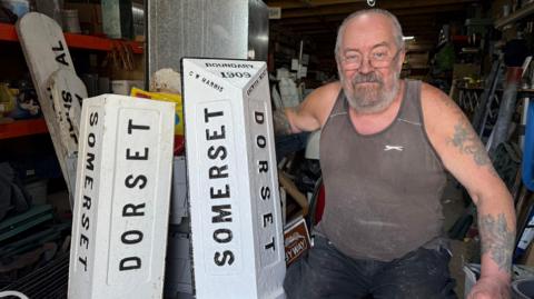 Two white signs on the floor, standing up, saying "Dorset" and "Somerset". There is a man with a beard crouching down next to them.