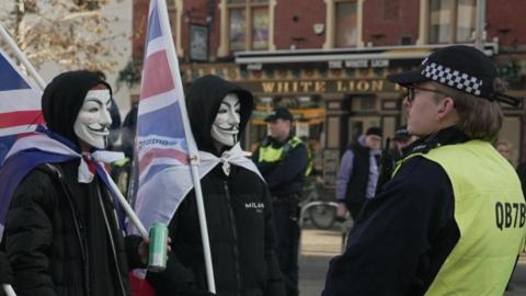 Three men wearing face masks in the style of the film V for Vendetta, white masks with black facial features, dressed in black and carrying Union flags, looking at two police officers facing them