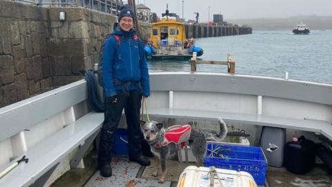 Laura Bambini is standing in the flat stern area of a boat that is tied up to the St Mary's harbour wall. She is holding Kuki's lead. Kuki is a grey and white dog with brown paws and she is wearing a red, grey and white coat. Laura is wearing navy coloured waterproofs, a hat and glasses. She is smiling. There are a couple of crates on the deck. There is a bright yellow boat tied up behind them and another boat under sail heading for the harbour. It is a grey day. 