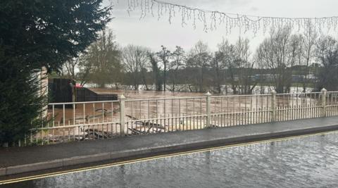 The image shows lots of water both on a road and what looks to be fields. The water on the fields is brown and seems to have risen high up the trees in the background.