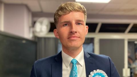 A head and shoulders shot of councillor Oliver Freeston smiling at the camera. He is wearing a blue blazer, a light blue, patterned tie, a white shirt and a light blue and white badge. He has short, blonde hair.
