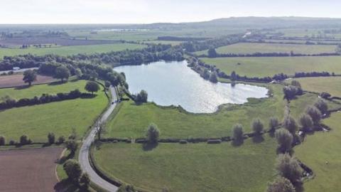 An aerial view of a lake amid green countryside, a road winding alongside.