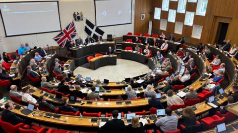 Cornwall Council chamber. People are sat on tables in chairs, in a circular shape. The chairs are red in colour. There are interactive boards displayed on the wall.