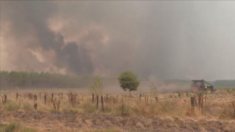 A tractor on the right hand side of the frame. Thick smoke fills the sky above farmland and forestry