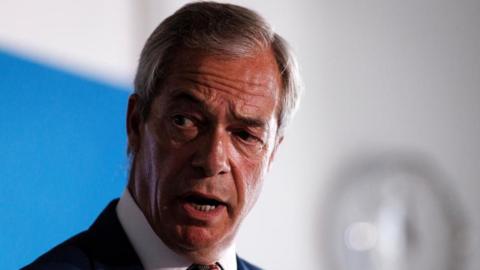 Headshot of Nigel Farage speaking a news conference. He is wearing a dark suit and multi-coloured tie.
