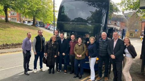 A group of people - 11 in total - stand in front of a double decker dark blue coach. It is parked on a hill.
