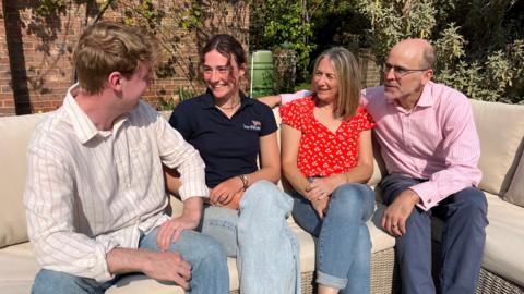 Zara Lachlan sitting with her boyfriend and parents on an outdoor sofa, while they talk amongst each other. Everyone is smiling. Zara has long brunette hair and is wearing a navy polo top. Boyfriend Isaac has short blonde hair and is wearing a white and pink striped shirt. Mother Claire has long blonde hair and is wearing a bright red top with white patterns. Father Guy has short black hair on the sides and back, and has glasses. He is also wearing a white and pink striped shirt, albeit different to Isaac's. All are wearing jeans.