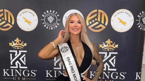 Victoria Emily, wearing a black dress and standing in front of a backdrop featuring a logo for Kings Gym. She is looking into the camera and smiling.