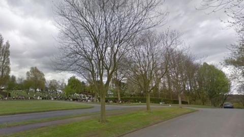 East Herringthorpe Cemetery in Rotherham, showing a drive with grass and trees on either side and gravestones in the background 