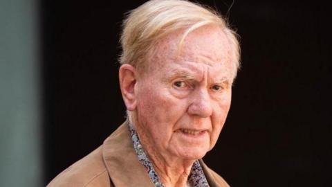 An older man in a pale brown jacket and paisley shirt looks toward the camera with a serious expression.