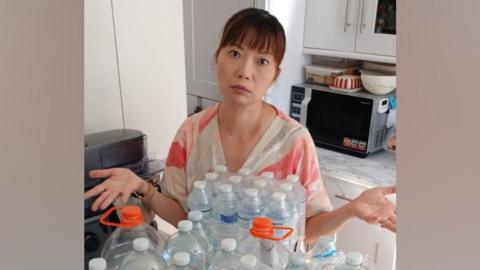 Pui Man is wearing a pink and white V-neck top. She has her hands raised by her sides - as if she is shrugging - and is pictured in her kitchen looking fed-up next to a pile of water bottles.