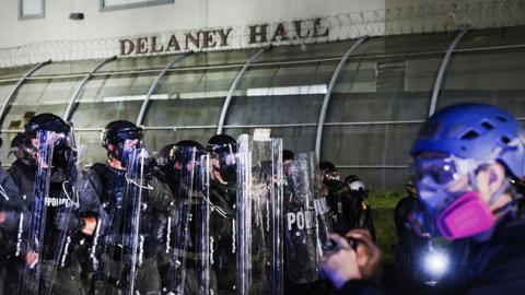 A line of police officers with riot shields form a line in front of the Delaney Hall immigration detention center in Newark, New Jersey. A person wearing a purple helmet and respirator is seen filming officers with a phone.