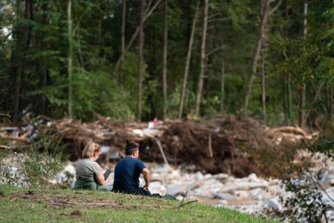 Two people sit on the edge of a hill overlooking damage from Hurricane Helene