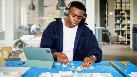 A young man wearing headphones using tablet and model to focus on architectural model design. 