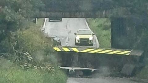 A road goes under a railway bridge, a section of which has collapsed. There are trees either side of the road. There is a yellow car on the far side of the bridge.