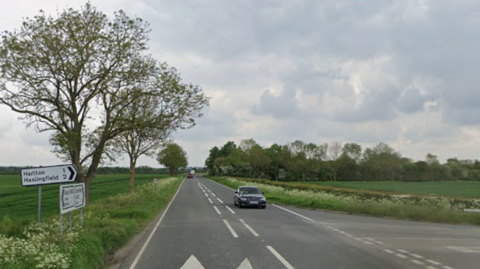 Street view of the A603, where the junction with Washpit Lane is to the right. To the left of the straight flat road are several road signs. One, pointing right, reads 'Harlton' and 'Haslingfield'. It is a cloudy day and there are several cars on the road. The road has green flat field on either side of it. 