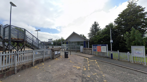Entrance area of Huntly railway station with a small station building, metal footbridge on the left, signage and noticeboards on the right, and trees in the background under a partly cloudy sky.