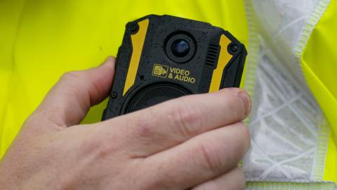 A close-up view of a hand holding a small black body‑worn camera. The device has yellow accents and raindrops on its surface. A circular lens sits near the top, and below it is a label showing an icon of a camera and the words "VIDEO & AUDIO". The person wearing it has a bright yellow high‑visibility jacket.